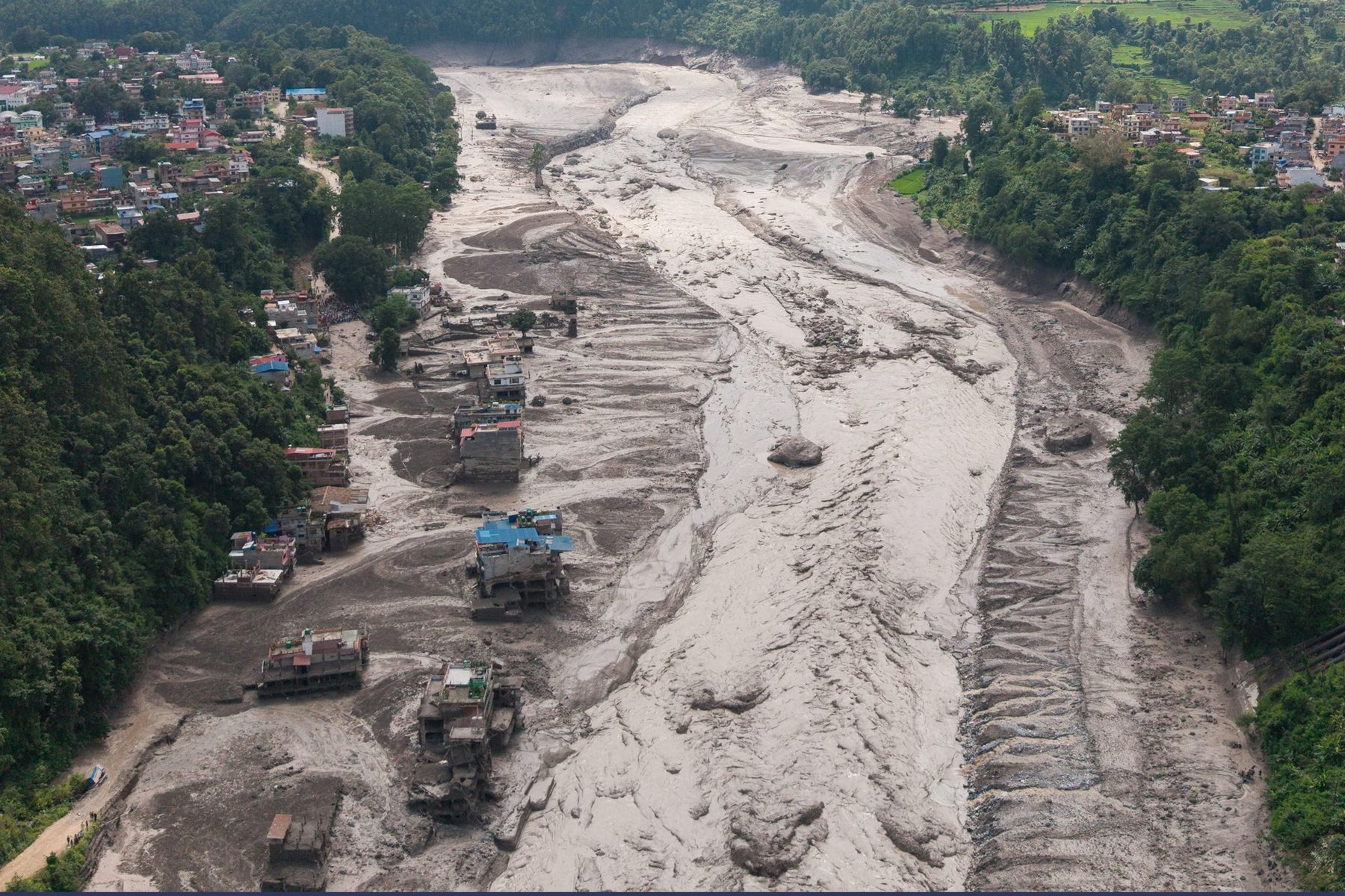 Aerial view of a river valley in Nepal filled with flood debris, with damaged buildings along its banks